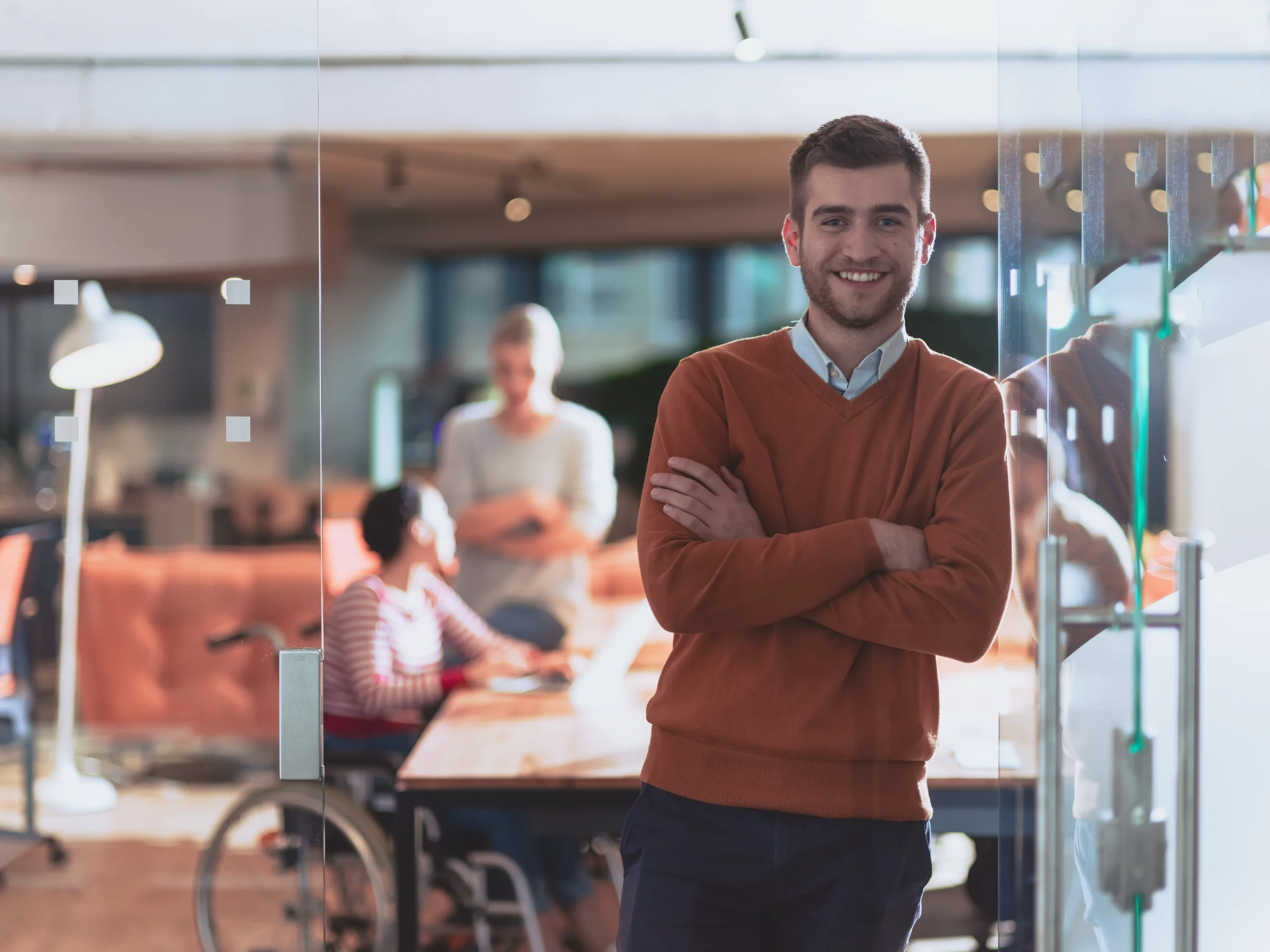 A young man in a brown sweater stands with his arms crossed, smiling in a modern insurance office. Behind him, people are working at a table, including a person in a wheelchair. The office has a contemporary, open-plan design.