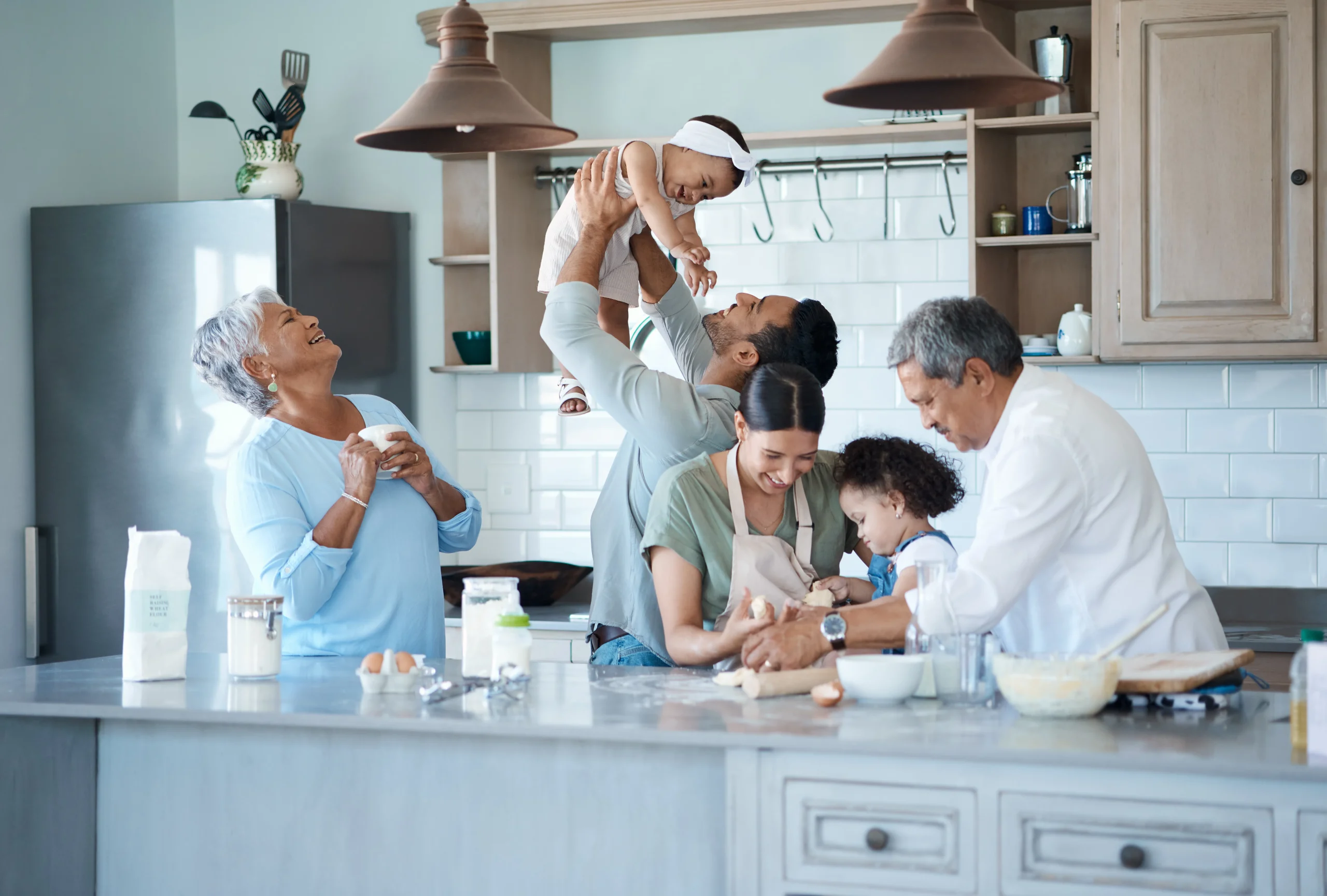 In a bright kitchen with modern decor, a joyful family scene unfolds: a man lifts a smiling baby while a woman and child bake together. Two older adults laugh nearby, embodying the warmth and security you cherish—just like comprehensive coverage to protect your home.