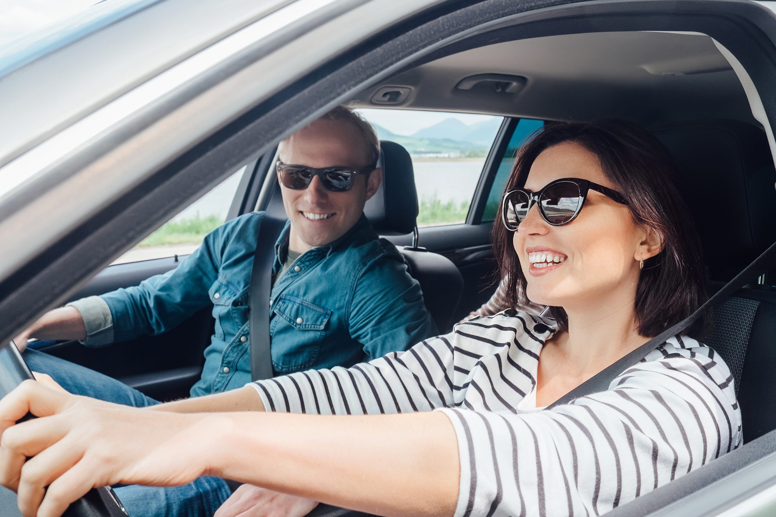 A man and a woman in sunglasses are smiling while sitting in a car, enjoying the drive. The woman is driving, both dressed casually. With a scenic view of mountains and a body of water behind them, it’s peace of mind knowing their adventure is protected by auto insurance.