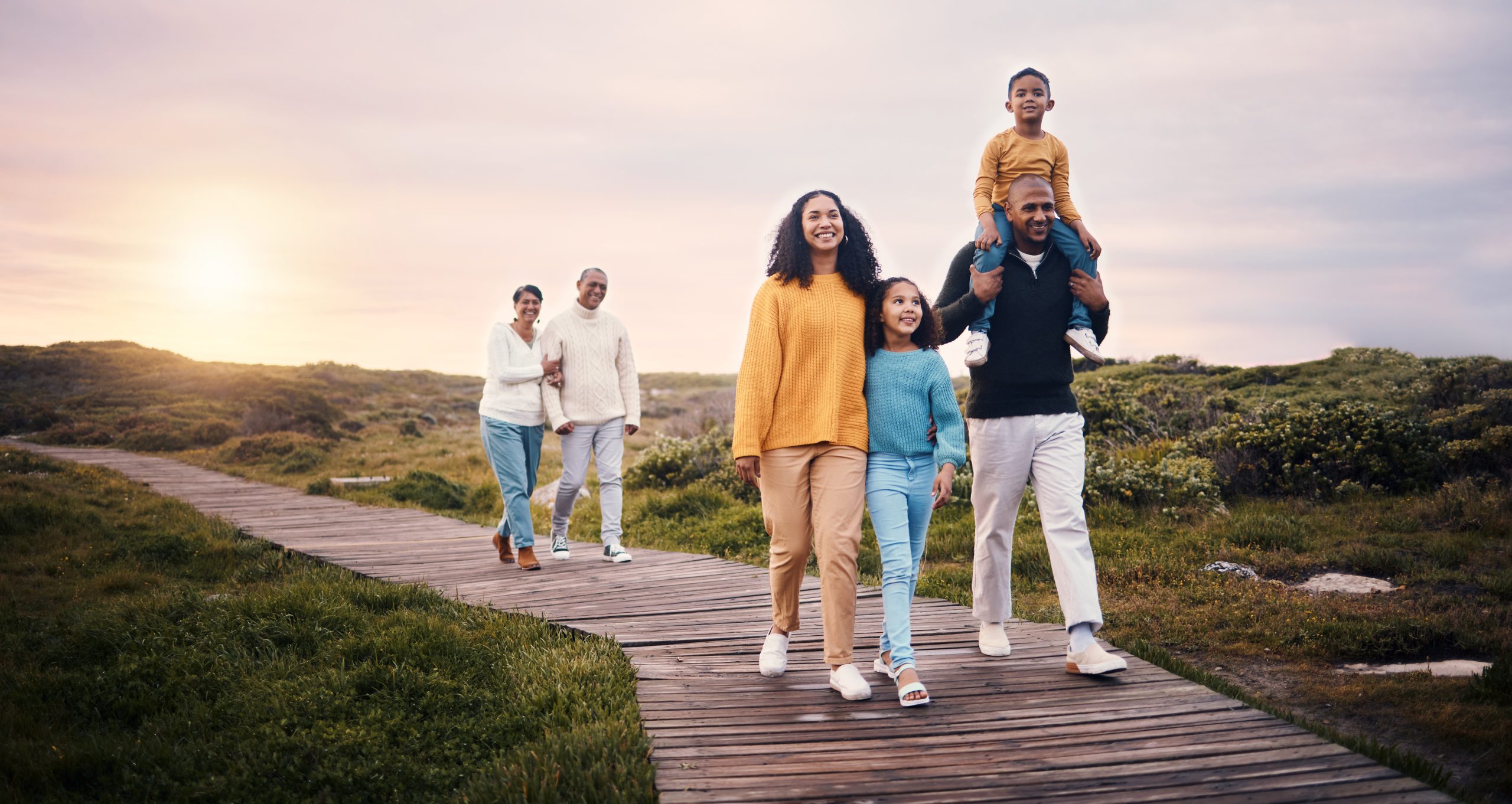 A diverse family strolls along a wooden path through a grassy landscape at sunset. The scene captures the joy of togetherness, as two adults lead with children in tow, one on a man's shoulders, while an older couple follows closely behind, all smiling and enjoying this cherished moment of security.