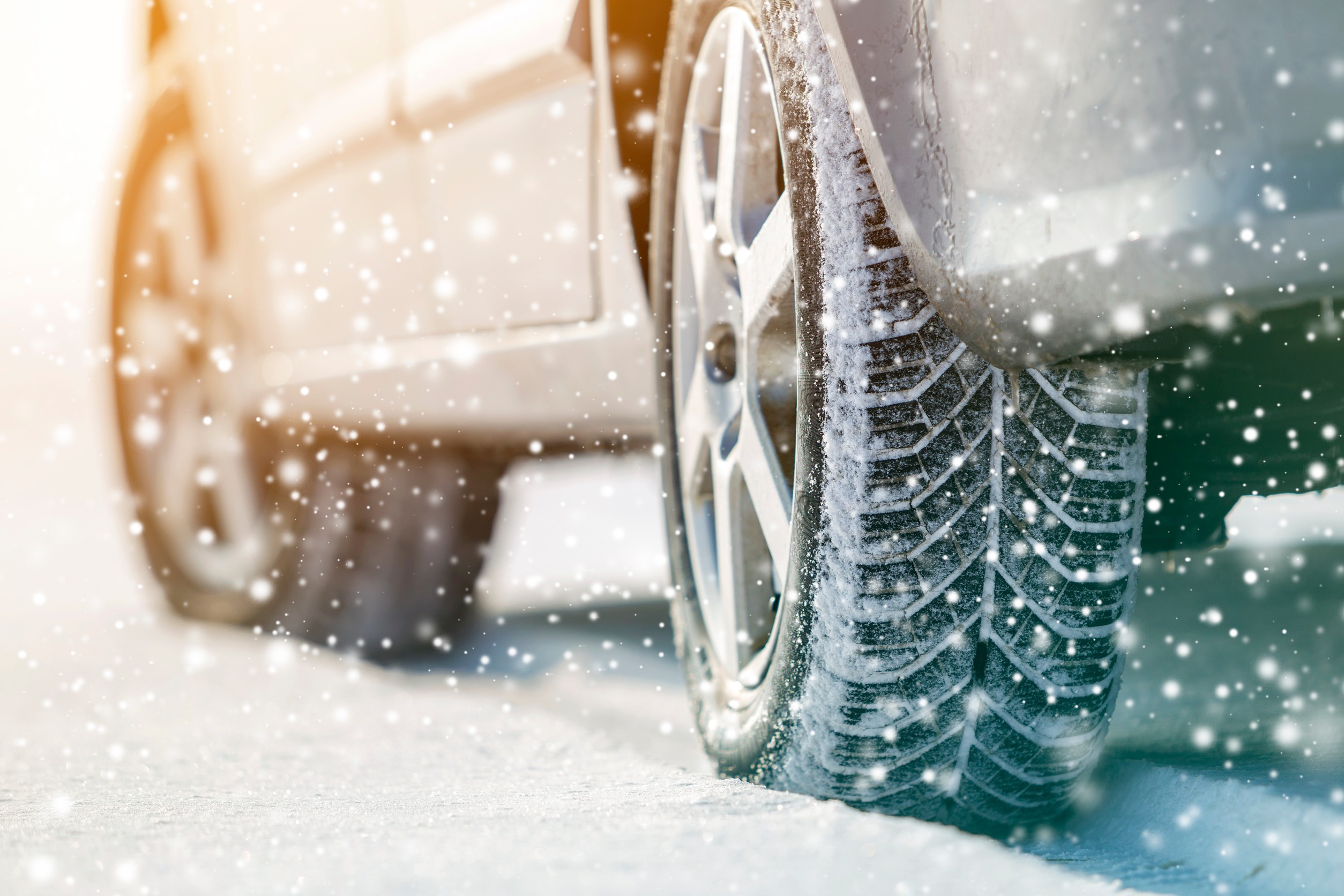 Close-up of a car's rear tire on a snowy Minnesota road. Snowflakes are falling, and the sunlight casts a warm glow, highlighting the tire's tread pattern essential for safe winter driving.