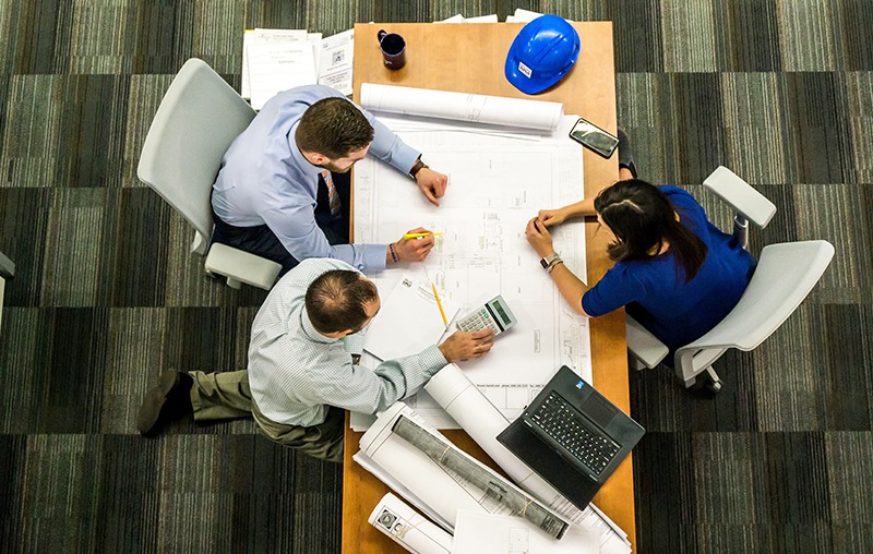 Three people are seated around a table covered with architectural blueprints. One person is using a calculator, another holds a pencil, and a laptop is open. A blue construction helmet and a smartphone are also on the table.