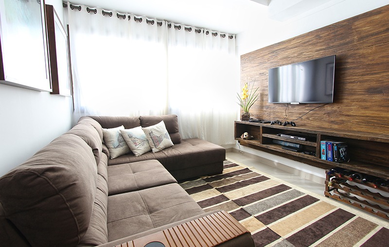 A cozy living room with a brown sectional sofa, decorative pillows, a wall-mounted TV, and a wooden entertainment unit. The room has a large window with curtains and a striped rug on the floor.