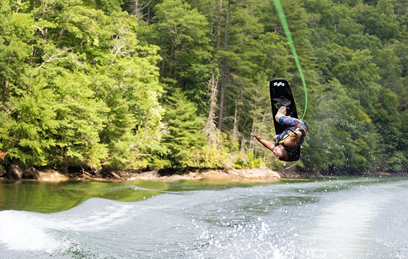 A person is performing an aerial flip while wakeboarding on a lake, surrounded by lush green trees. The water surface is disturbed by the boat's wake, and the sky is clear.