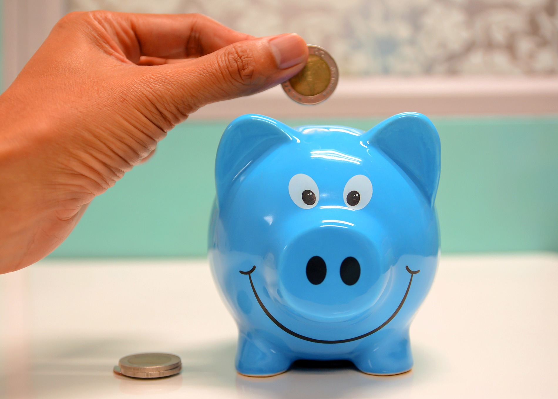 A person is inserting a coin into a smiling blue piggy bank on a white surface. Additional coins are placed next to the piggy bank. The background is softly blurred, giving a cheerful atmosphere.