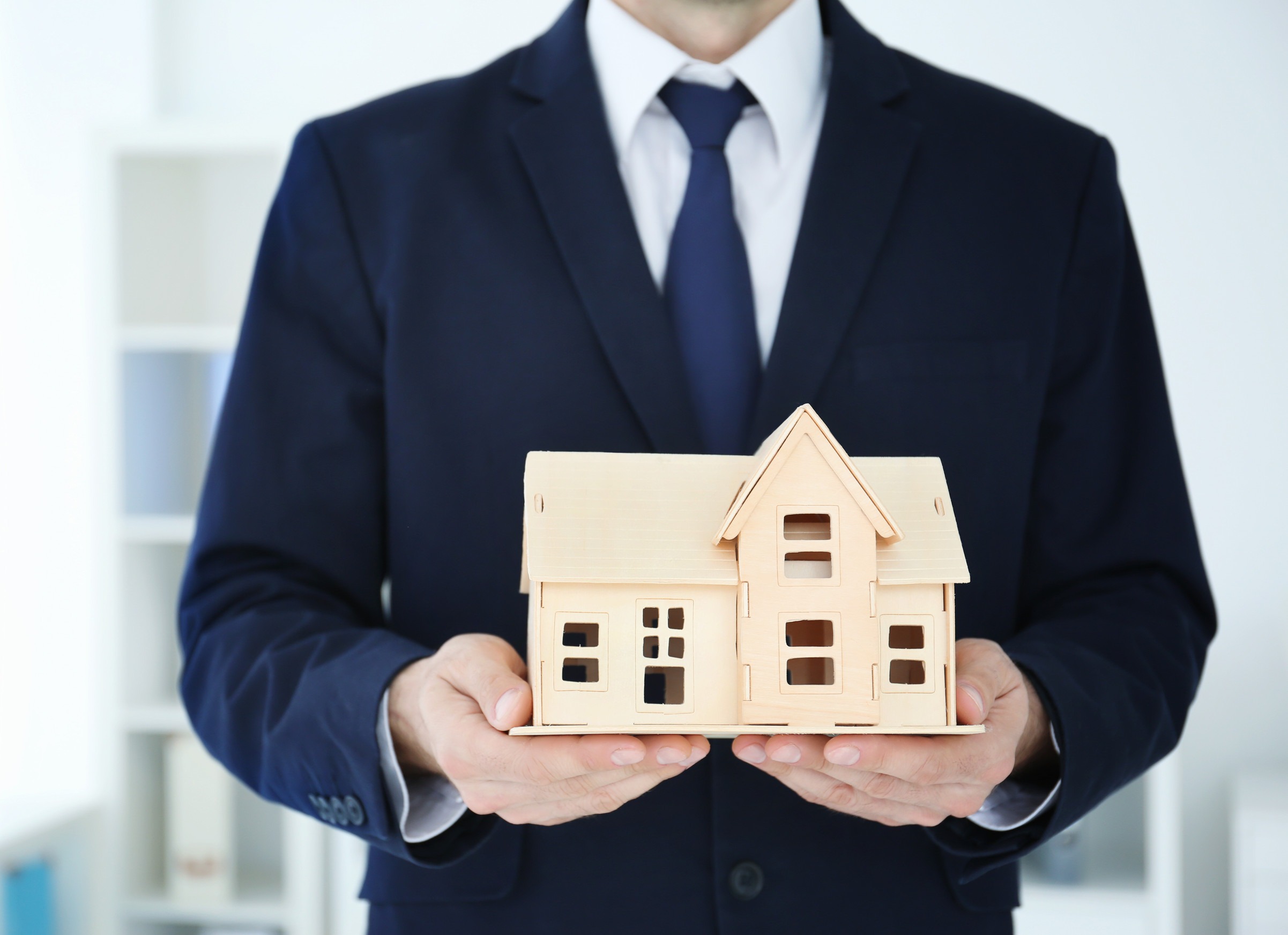 A person in a suit holds a small wooden house model in front of them. The background is softly blurred, suggesting an indoor setting.