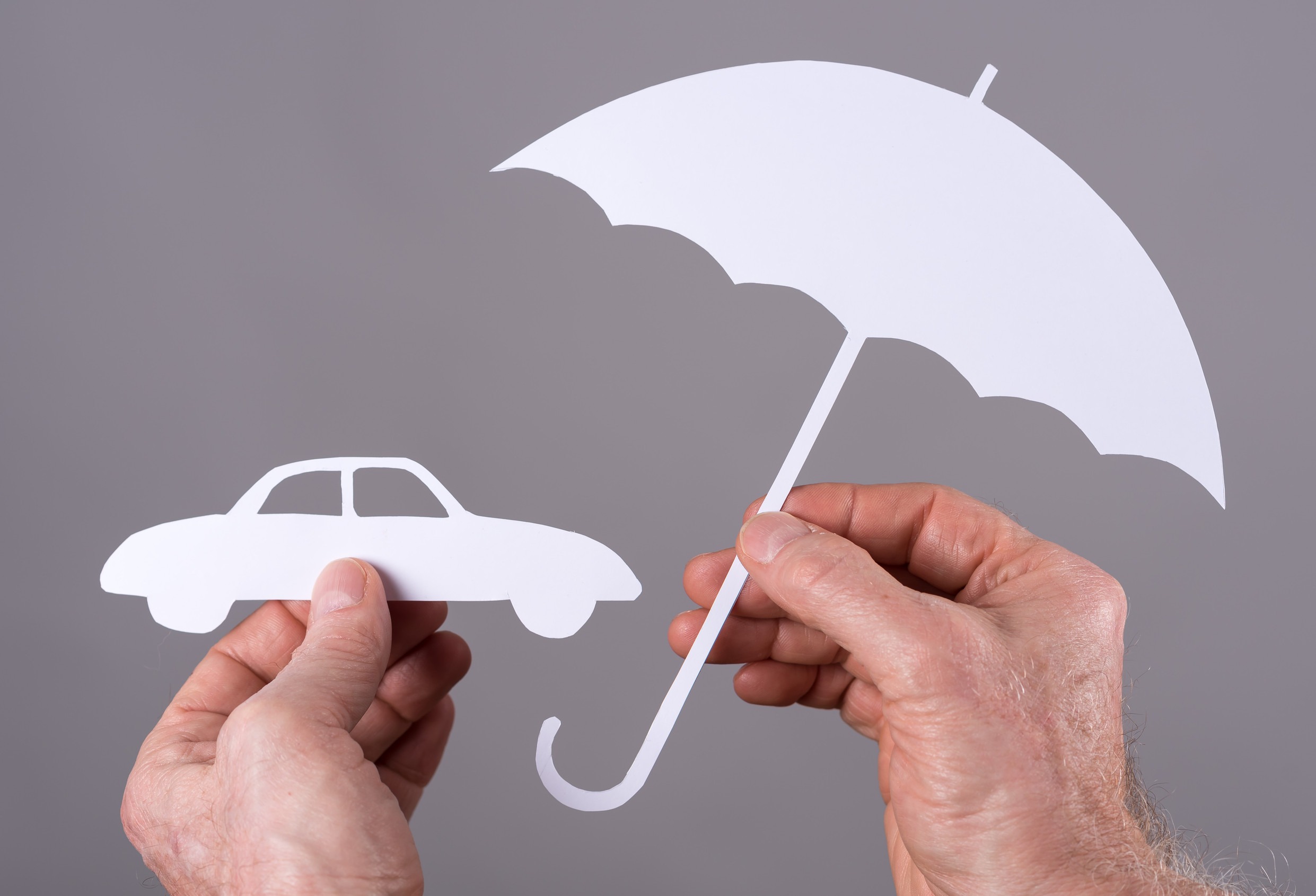 A person holds a paper cutout of a car in one hand and a white umbrella in the other, symbolizing coverage and protection for vehicles, highlighting auto insurance needs against a gray background.