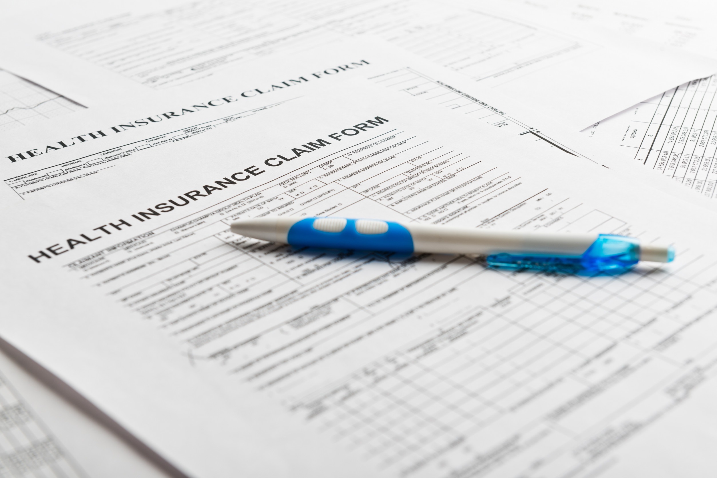 A blue and white pen rests on top of a stack of insurance claim forms, addressing a headache-inducing array of printed text. These forms include sections for personal and medical information, meticulously spread out across a light surface.