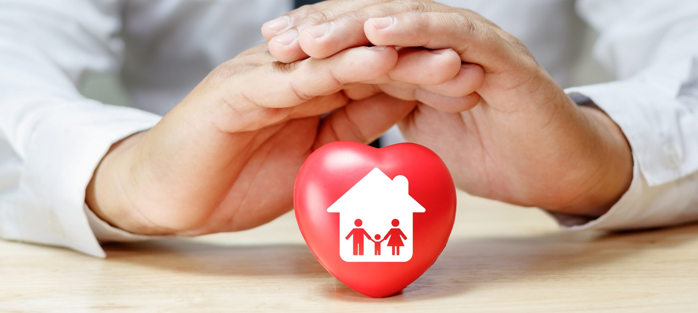 A person in a white shirt and black tie gently cups their hands over a red heart-shaped object on a table. The heart features a white graphic of a house with a family symbol inside it.