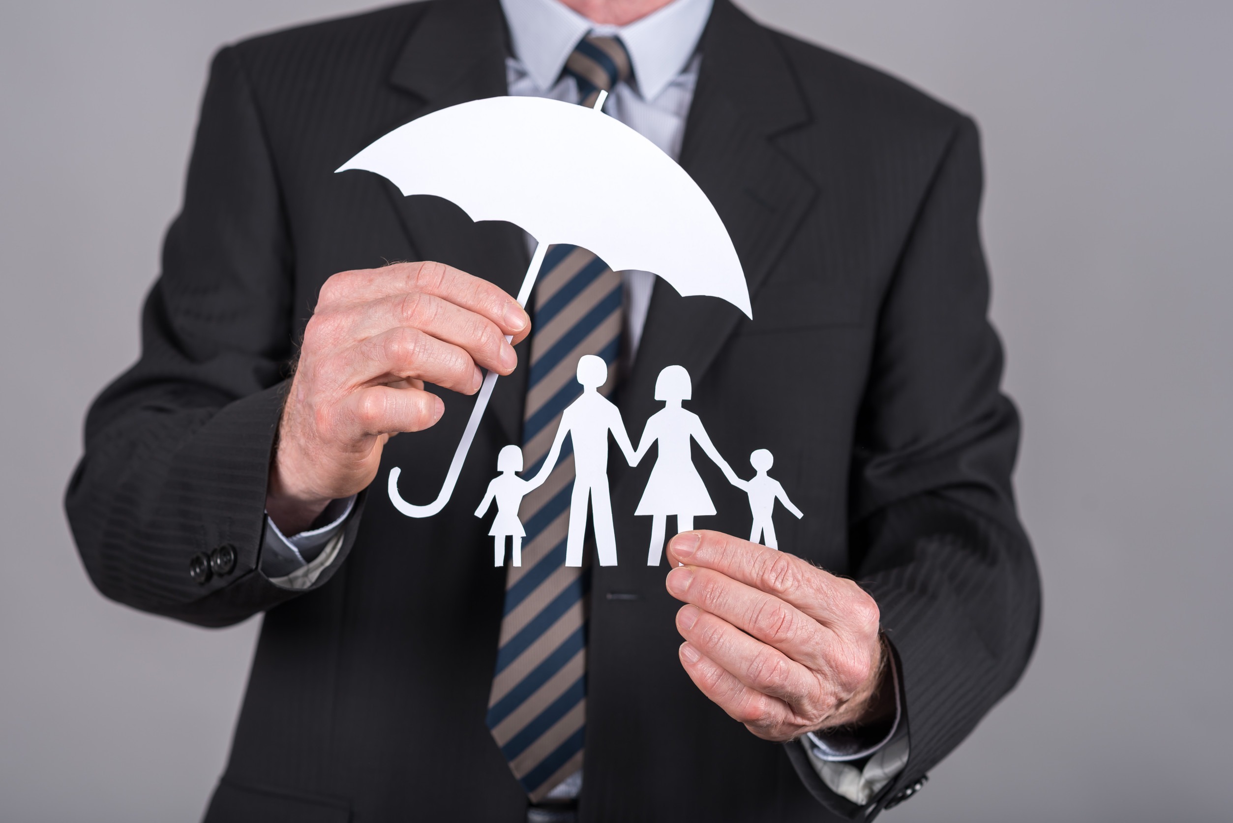 A person in a suit holds a paper cutout of a family under an umbrella, symbolizing umbrella insurance. The cutout depicts a man, woman, and two children holding hands, embodying the smartest coverage needed for protection and safety.