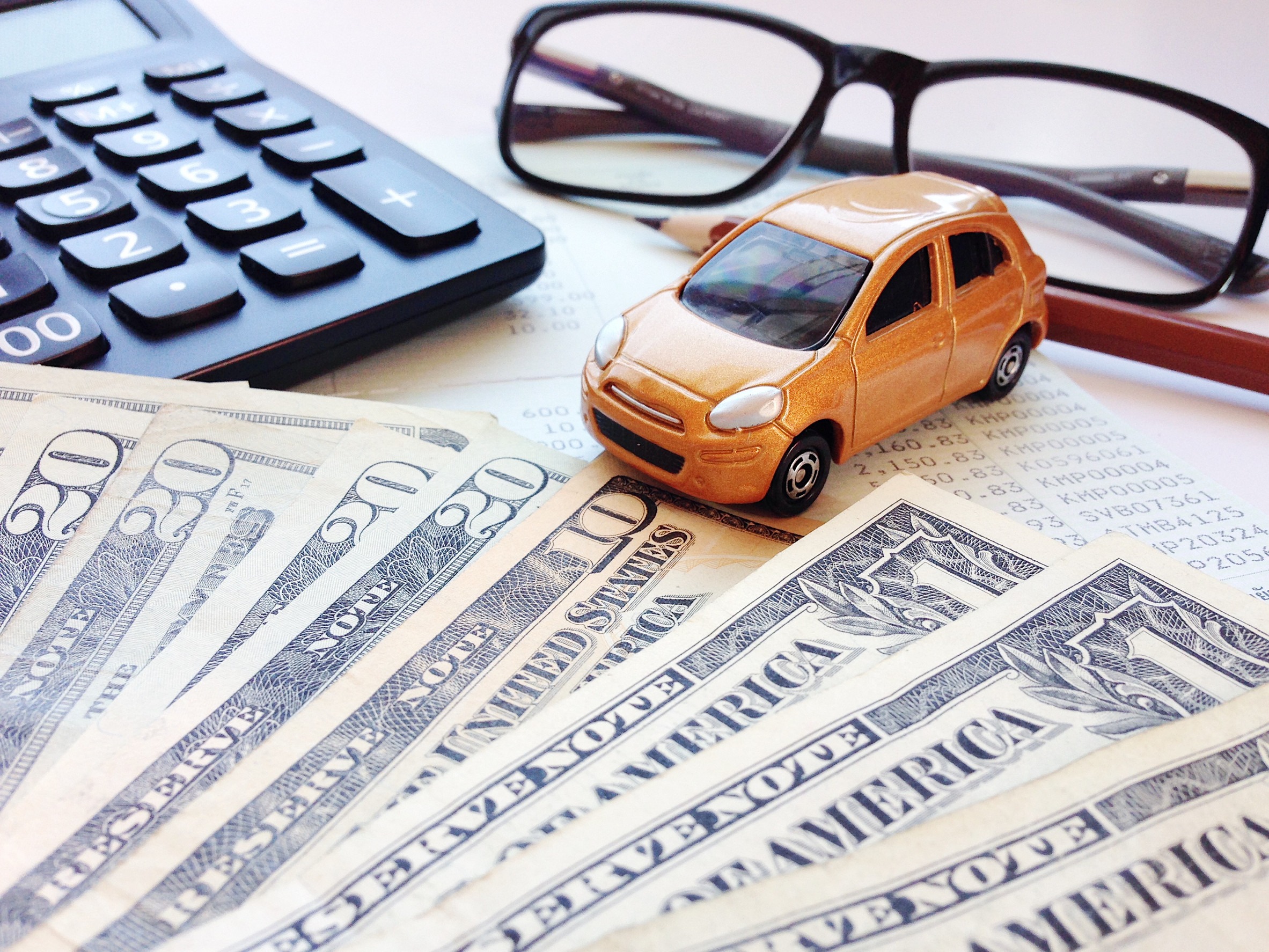 A small toy car rests on printed financial documents alongside a calculator. Nearby, a fan of U.S. bills is spread out, hinting at budget planning and car expenses. Glasses lie in the corner, emphasizing the importance of coverage considerations in managing a lower insurance bill.