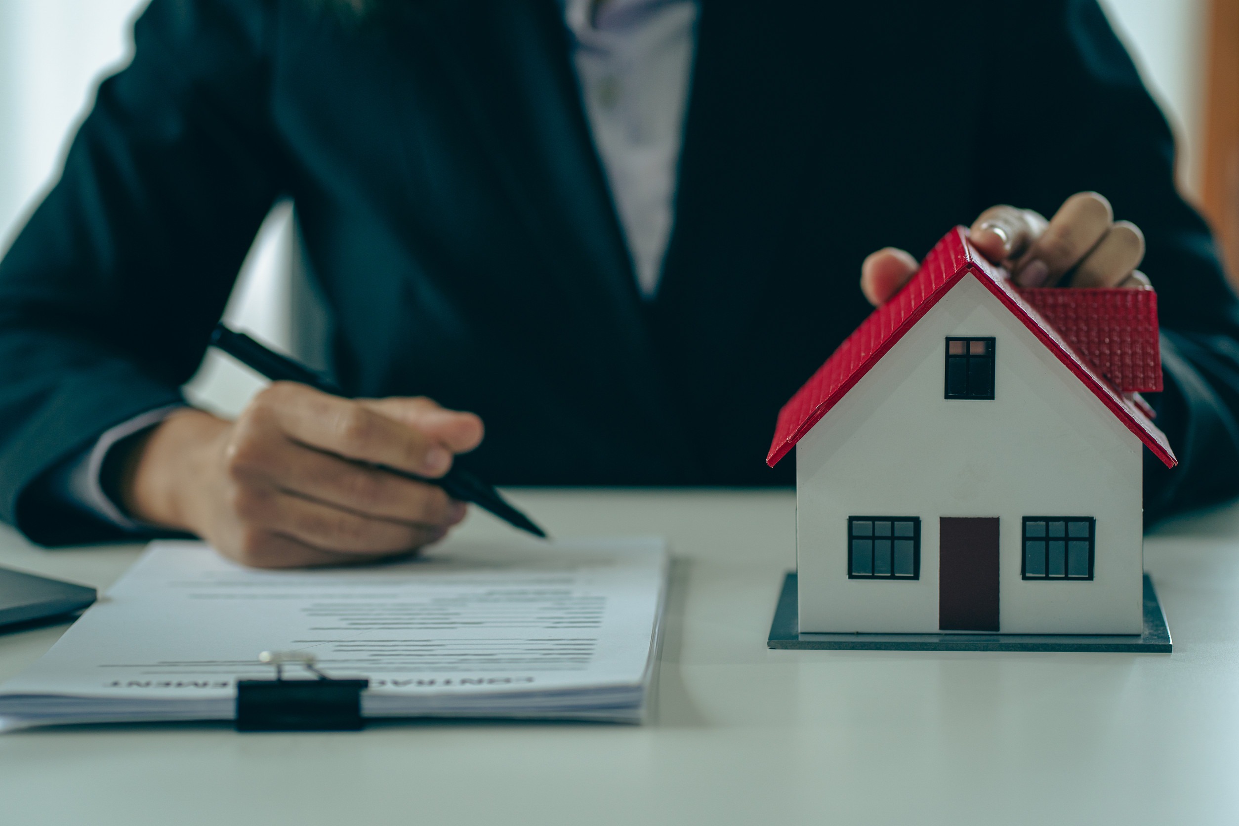 A person in a suit sits at a desk, holding a pen in one hand and a small model of a house with a red roof in the other. Papers are laid out on the desk, suggesting a real estate or contractual setting.