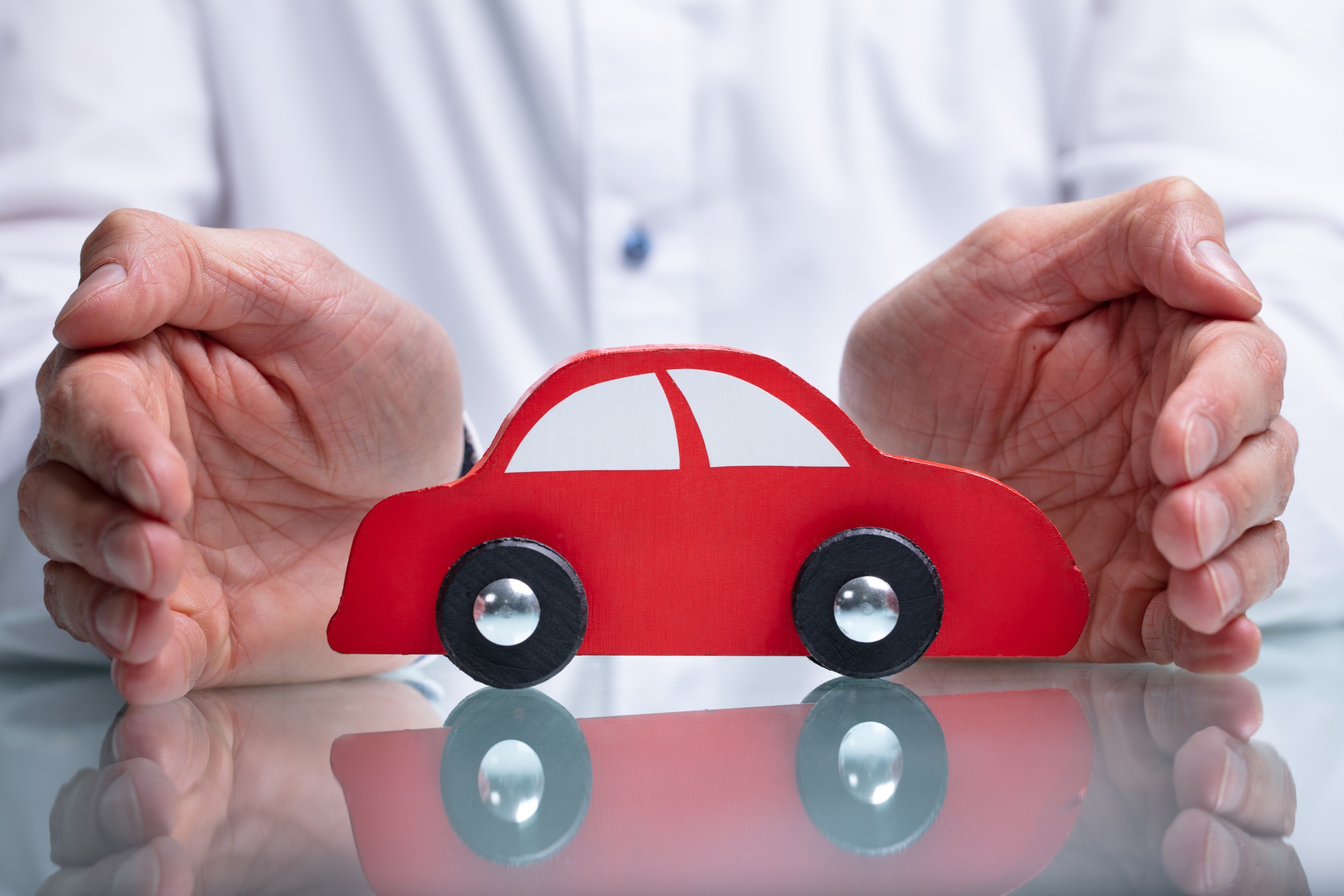 Hands gently cupping a small red toy car on a table, evoking a sense of protection—much like choosing the right auto insurance. The simple, stylized car with black wheels rests securely as the person in a white shirt ensures no overpaying for peace of mind.