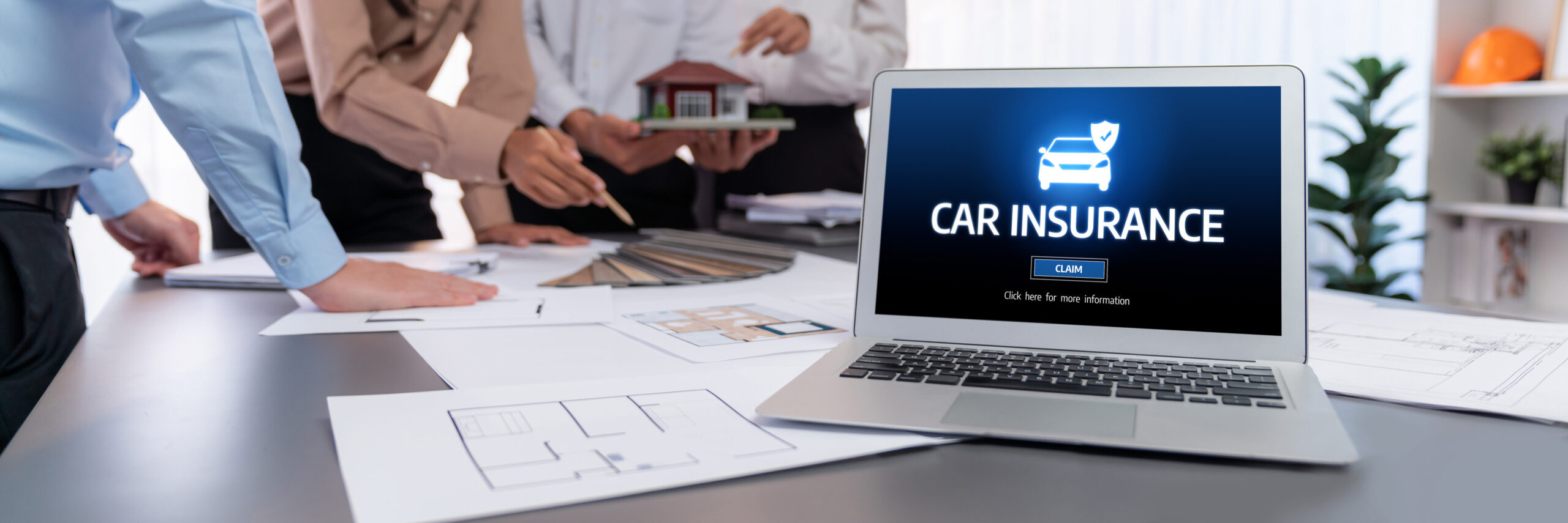 A laptop displaying a car insurance website sits on a table with architectural plans, while three people in business attire discuss documents and a model house—highlighting how to choose auto insurance that provides the right coverage.