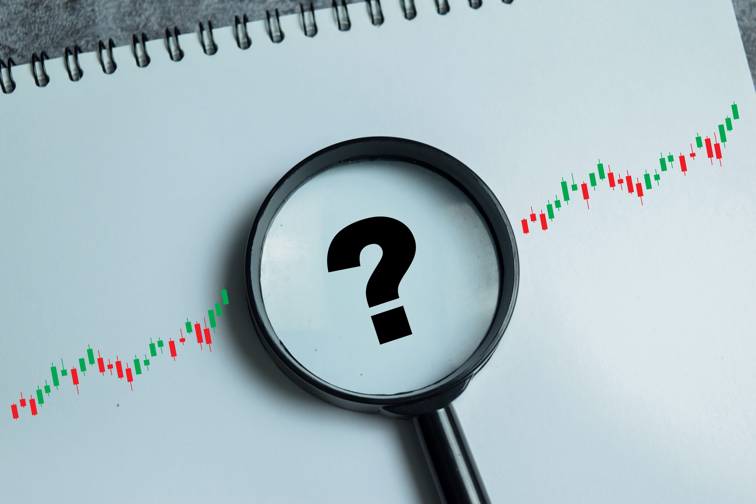 A magnifying glass with a question mark inside is placed over a rising candlestick chart on a spiral notebook, symbolizing financial analysis or uncertainty in stock market trends and the reasons for premium increase in insurance rates.